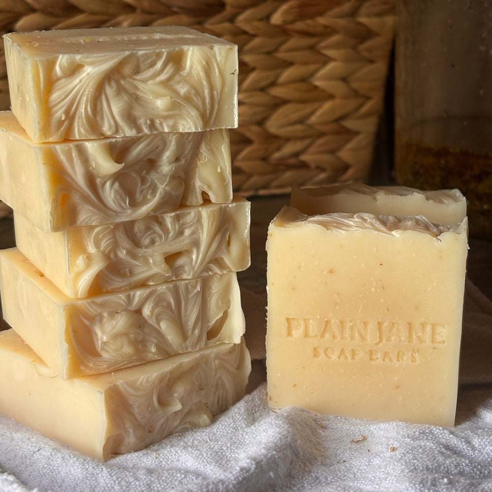 Five handmade soap bars with a swirled marbled pattern are stacked on the left, next to one bar embossed with 'PLAIN JANE SOAP BAR' on the right, set on a white cloth over a rustic wooden surface with a woven basket in the background.
