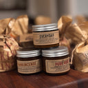 Three small glass jars with silver lids and beige labels reading Everyday, Frankincense, and Pure Rose, arranged on a wooden surface with brown paper bags in the background.