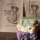 Three bars of soap in green, white, and purple on a rustic wooden surface with 'Plain Jane Soap Bars' packaging.